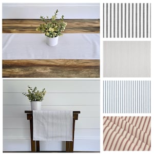 May include: A rectangular table runner with blue and white stripes is displayed on a wooden table. A small white pot with greenery sits on the table. Additional fabric swatches with various stripe patterns are shown.