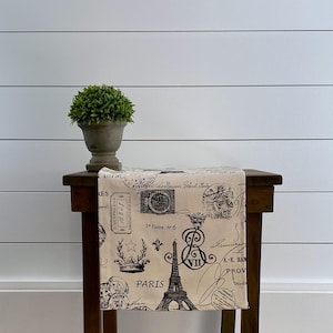 May include: A beige table runner with a black and white Parisian themed print. The runner features images of the Eiffel Tower, stamps, and French script. The runner is on a wooden table with a small potted plant on the side.