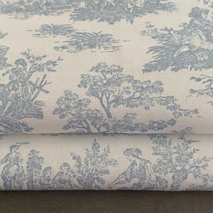 May include: Close-up of a folded fabric featuring a light blue toile de Jouy pattern on a white background. The design depicts pastoral scenes with figures, trees, and architectural elements, creating a classic, elegant aesthetic.