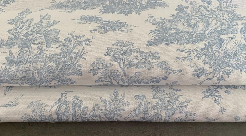 May include: Close-up of a folded fabric featuring a light blue toile de Jouy pattern on a white background. The design depicts pastoral scenes with figures, trees, and architectural elements, creating a classic, elegant aesthetic.
