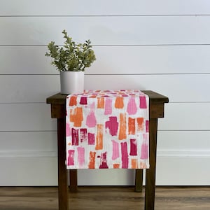 May include: A white table runner with a pink, orange, and white abstract brushstroke pattern. The runner is on a wooden table with a potted plant on top.