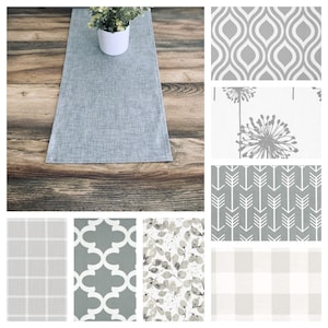 May include: A gray table runner with a subtle texture, displayed on a wooden table. The runner is surrounded by eight different patterns in gray and white, including geometric, floral, and plaid designs.
