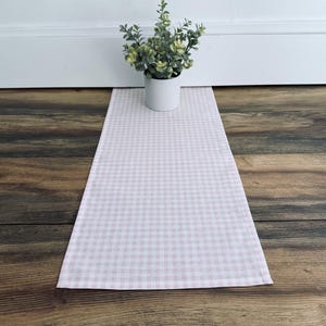 May include: A pink and white gingham patterned table runner. A small potted plant sits on the runner. The runner is rectangular and made of fabric. The background is a wooden floor and a white wall.