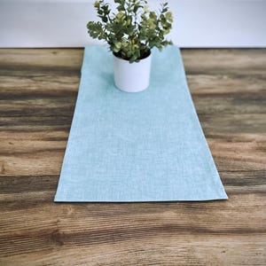 May include: A light blue table runner with a textured appearance, placed on a wooden surface. A small white pot with a green plant sits on the runner. The runner is rectangular and appears to be made of fabric.