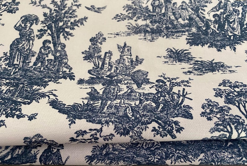 May include: Cream-colored fabric with a repeating toile pattern in navy blue. The design depicts pastoral scenes with figures, trees, and architectural elements. The fabric is folded, showing the pattern's continuity.