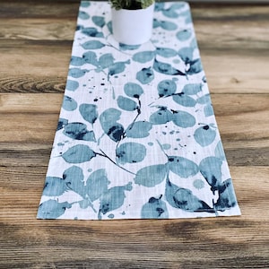 May include: A white table runner with a blue and white watercolor leaf pattern. The runner is on a wooden table.