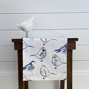 May include: A white table runner with a blue and gray bird pattern. The runner is on a wooden table with a white ceramic bird figurine on top.