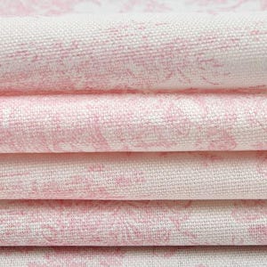 May include: Close-up of folded fabric with a pink floral pattern on a white background. The fabric is stacked in layers, showcasing the texture and design. The pink pattern is a soft, watercolor-like design.