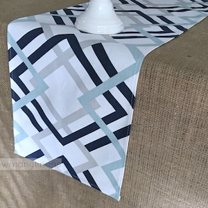 May include: A white table runner with a geometric pattern in shades of blue and grey. The pattern features intersecting lines and squares. The runner is shown on a hessian surface.