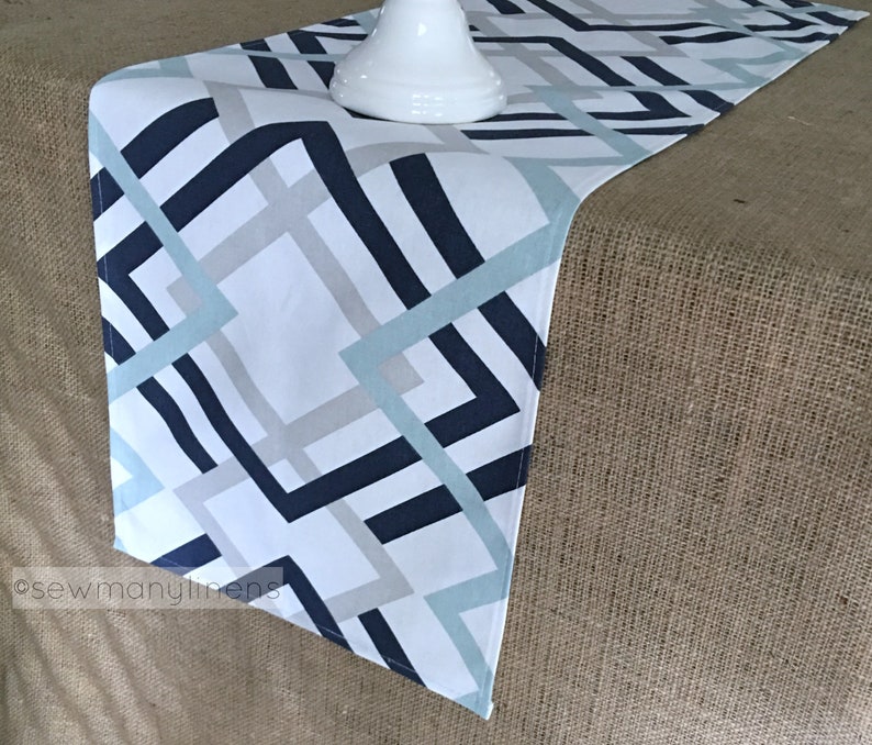 May include: A white table runner with a geometric pattern in shades of blue and grey. The pattern features intersecting lines and squares. The runner is shown on a hessian surface.