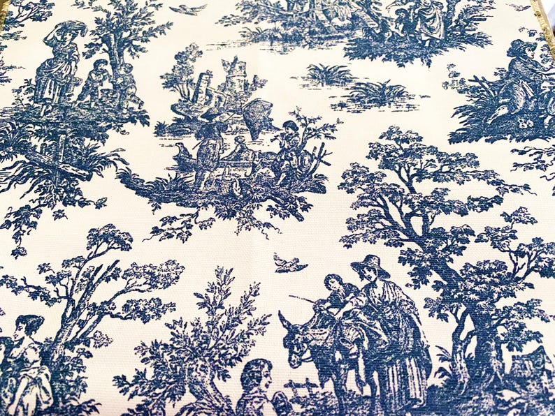 May include: Cream-colored fabric with a repeating blue toile pattern. The design depicts pastoral scenes with figures, trees, and buildings. The overall aesthetic is reminiscent of classic French country decor.