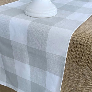 May include: A white and gray checkered table runner. The runner is made of a soft, woven fabric and has a subtle sheen. It is laid on a brown burlap surface.