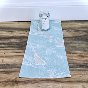 May include: A small glass jar with a metal handle and a white substance inside sits on a light blue placemat. The placemat features a nautical design with white sailboats and waves.