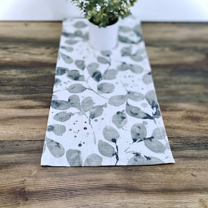 May include: A white table runner with a gray watercolor leaf pattern. The runner is on a wooden table.
