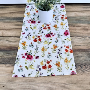 May include: A rectangular floral table runner with a white background. The runner is decorated with a pattern of red, pink, yellow, and purple flowers and green stems. A small white pot with a green plant sits on top of the runner.
