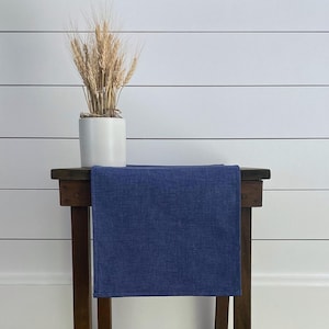 May include: A blue table runner on a wooden table with a white vase and dried wheat stalks.
