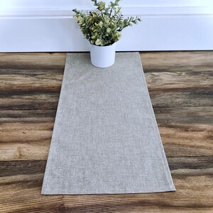 May include: A light gray linen table runner with a subtle texture. The runner is laid out on a wood floor.