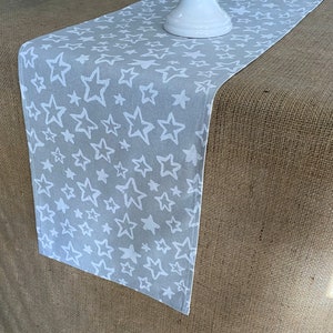 May include: A gray table runner with a white star pattern. The runner is on a brown burlap surface.