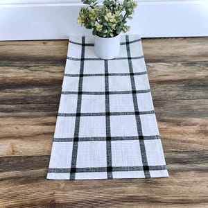 May include: A white table runner with a black plaid pattern. A small white pot with a green plant sits on the runner. The runner is made of a woven fabric and is rectangular in shape.
