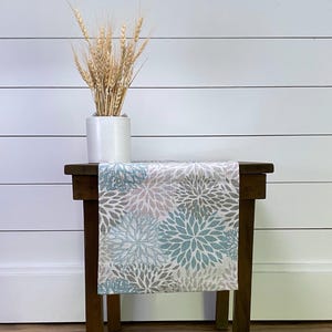May include: A light blue and brown floral table runner with a white background. The runner is on a wooden table with a white vase and wheat stalks.