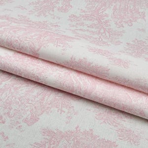 May include: Close-up of a folded fabric with a pink toile de Jouy pattern on a white background. The design features detailed illustrations of trees and landscapes. The fabric has a soft, textured appearance.
