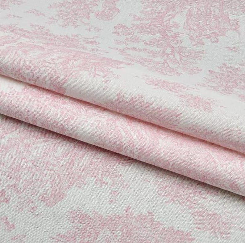 May include: Close-up of a folded fabric with a pink toile de Jouy pattern on a white background. The design features detailed illustrations of trees and landscapes. The fabric has a soft, textured appearance.