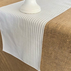 May include: White table runner with thin gray stripes. The runner is laying on a brown burlap surface.