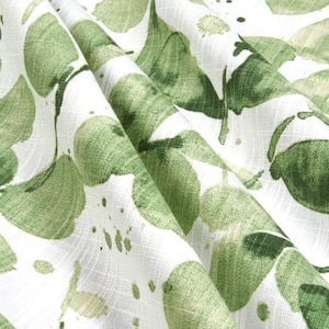 May include: Close-up of white fabric with a green leaf pattern. The leaves are large and abstract, with a watercolor-like effect. The fabric is folded, showing the texture and depth of the material. The design is reminiscent of nature and botanical themes.