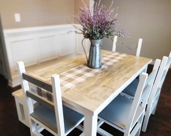 May include: A white farmhouse style dining table with four chairs. The table has a wood top and a white base. The chairs are also white with grey cushions. A checkered tablecloth is on the table with a metal pitcher filled with purple flowers.
