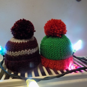 May include: Two knitted hats with pom-poms, one red and green, the other burgundy and white, are sitting on a white radiator with a string of Christmas lights.