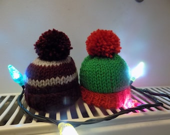 May include: Two knitted hats with pom-poms, one red and green, the other burgundy and white, are sitting on a white radiator with a string of Christmas lights.