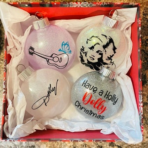 May include: Four silver glitter Christmas ornaments with different designs. One ornament has a guitar and butterfly, one has a portrait of Dolly Parton, one has a signature that says "Dolly", and one says "Have a Holly Dolly Christmas".