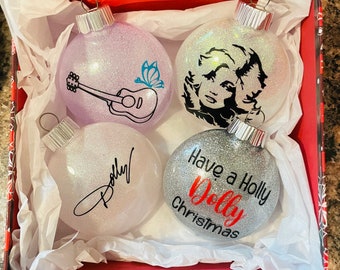 Puede incluir: Cuatro adornos navideños redondos y brillantes en una caja de regalo. Uno es morado claro con una guitarra negra y una mariposa azul. Uno es transparente con un retrato negro. Uno es transparente con el nombre Dolly. Uno es plateado con texto.