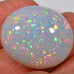 Puede incluir: Una piedra preciosa de ópalo pulida, de forma ovalada. El ópalo tiene un color base blanco lechoso con destellos iridiscentes de verde, naranja, amarillo y azul. La superficie de la piedra es lisa y reflectante, mostrando un juego de colores.