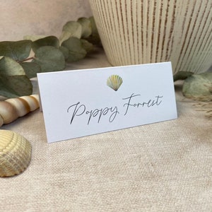 May include: White place card with a small seashell illustration and the name "Poppy Forrest" written in black cursive.