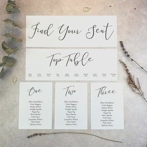 May include: White cards with black calligraphy text for a wedding reception seating chart. The top card says "Find Your Seat". The next card says "Top Table". The remaining cards are numbered "One", "Two", and "Three" and list names for each table.
