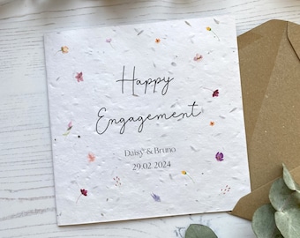 May include: A white card with the text "Happy Engagement" written in black cursive. Below the text is "Daisy & Bruno" and "29.02.2024". The card is decorated with small, colorful flowers.