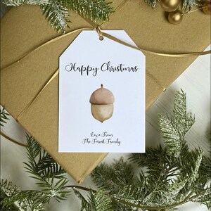 Op de afbeelding: Witte cadeau-tag met de tekst "Happy Christmas" in zwarte cursieve letters. Een eikel-illustratie bevindt zich in het midden van de tag. De tekst "Love from The Forrest Family" staat onderaan de tag.