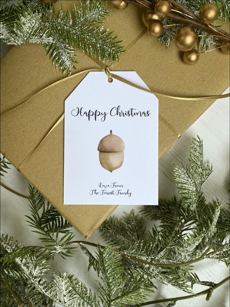 Op de afbeelding: Witte cadeau-tag met de tekst "Happy Christmas" in zwarte cursieve letters. Een eikel-illustratie bevindt zich in het midden van de tag. De tekst "Love from The Forrest Family" staat onderaan de tag.