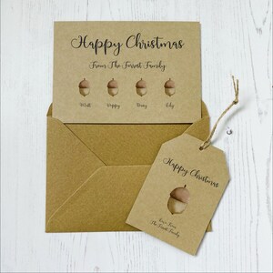Op de afbeelding: Een bruine kaart met de tekst "Prettige Kerst" en "Van de familie Forrest". Er zijn vier eikels met de namen "Matt", "Poppy", "Daisy" en "Lily" eronder geschreven. Een bruine envelop en een bruin cadeau-etiket met de tekst "Prettige Kerst" en "Lieve groeten van de familie Forrest" zijn ook te zien.