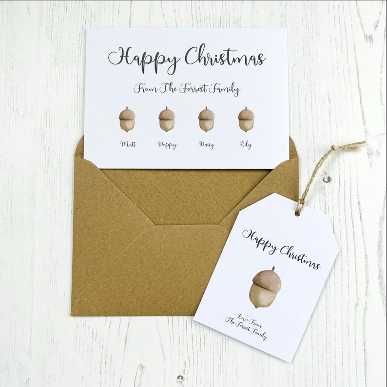 Op de afbeelding: Een witte kaart met de tekst "Prettige Kerst" en "Van de familie Forrest" in zwarte cursieve letters. Er zijn vier eikel-illustraties met de namen "Matt", "Poppy", "Daisy" en "Lily" eronder geschreven. Een bruine envelop en een wit cadeau-etiket met de tekst "Prettige Kerst" en "Lieve groeten van de familie Forrest" zijn ook inbegrepen.