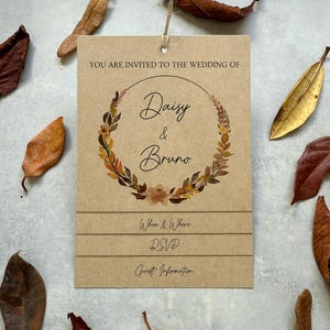 May include: A brown card wedding invitation with the text "YOU ARE INVITED TO THE WEDDING OF Daisy & Bruno". The card features a floral wreath design and is surrounded by dried leaves. A twine string is attached to the top of the card.
