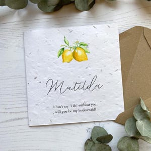 May include: A white invitation card with the name "Matilda" written in cursive. It features a watercolor illustration of two yellow lemons with green leaves and white flowers. The text reads, "I can't say 'I do' without you, will you be my bridesmaid?"