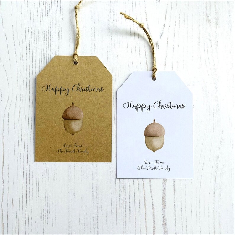 Op de afbeelding: Twee cadeaulabels met een bruine eikel-illustratie en de tekst "Prettige Kerst" en "Liefde van de familie Forrest". Een label is bruin en de andere is wit.