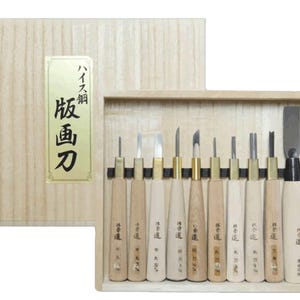 May include: A set of eleven wood carving tools with light-colored wooden handles and metal blades, arranged in a light wood storage box. The box lid features a gold label with black Japanese text. The tools vary in blade width and shape.