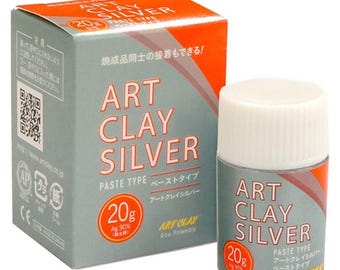 Pode incluir: Pasta de argila prateada num pequeno recipiente cilíndrico com tampa branca, ao lado de uma caixa a condizer. A caixa e o recipiente apresentam o texto "ART CLAY SILVER" em letras grandes, com texto japonês por baixo. O recipiente contém 20g de argila.