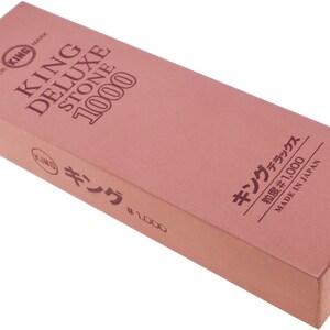 May include: A rectangular pink sharpening stone with the text KING DELUXE STONE 1000 printed on the top surface. The side features the text KING, Japanese characters, and #1000. Additional Japanese text and MADE IN JAPAN are printed on the side.