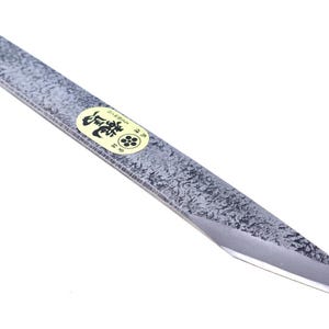 Pode incluir: Uma faca utilitária de gume único com um acabamento de metal martelado cinzento-escuro texturizado na lâmina e no cabo. Um pequeno autocolante circular amarelo com caracteres japoneses pretos e um emblema floral está colado no centro da lâmina.