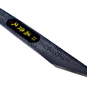 Pode incluir: Uma faca japonesa kiridashi de gume único com um acabamento de metal escuro, texturizado e martelado na lâmina. A lâmina inclui uma incrustação oval preta com caracteres kanji japoneses amarelos e uma ponta afiada para cortes de precisão.