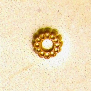 May include: A gold-colored metal button with a circular design. The button has a raised center and a ring of small, evenly spaced dots around the edge.